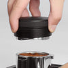 A hand is holding a black precision espresso distribution tool above a metal container filled with brown grounds.