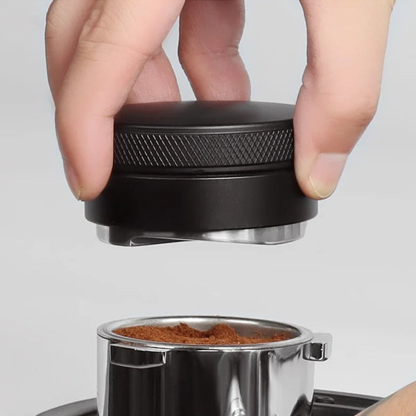 Eine Hand hält ein schwarzes Präzisions-Espresso-Verteilungswerkzeug über einen Metallbehälter mit braunen Kaffeebohnen.