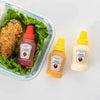 A glass container holds a fried chicken piece and three mini squeeze bottles of tomato ketchup, honey mustard, and honey must