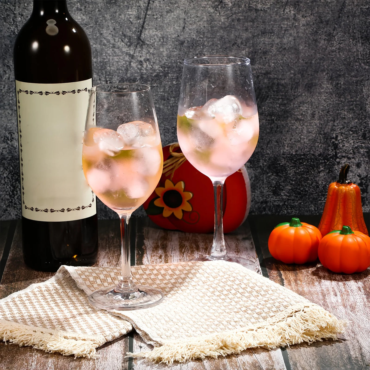 A glass of pink cocktail with ice cubes sits in front of a bottle and orange decorative items.