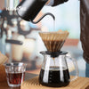 A black pour over coffee pot is pouring coffee into a glass, set on a wooden surface.