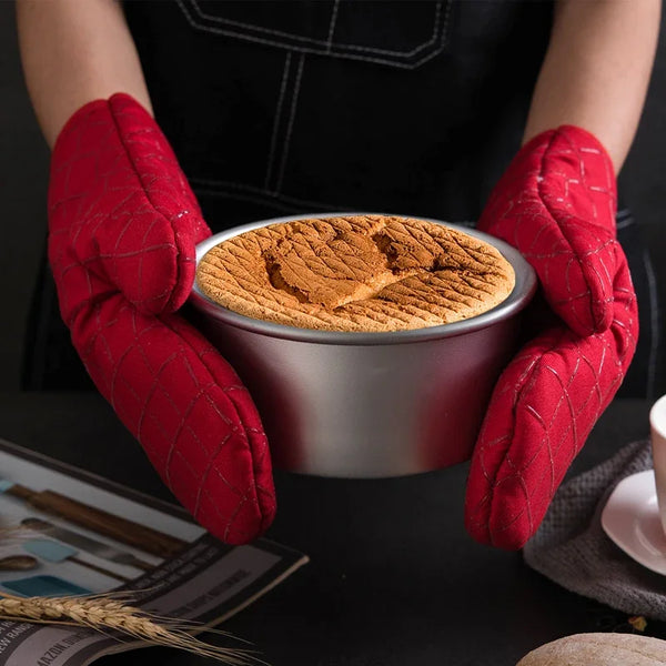 A pair of red silicone printed non-slip oven mitts are shown holding a round pan filled with brown, textured food.