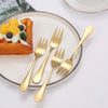 A set of six gold-toned stainless steel dessert forks is displayed next to a plate with a piece of orange waffle and cream.