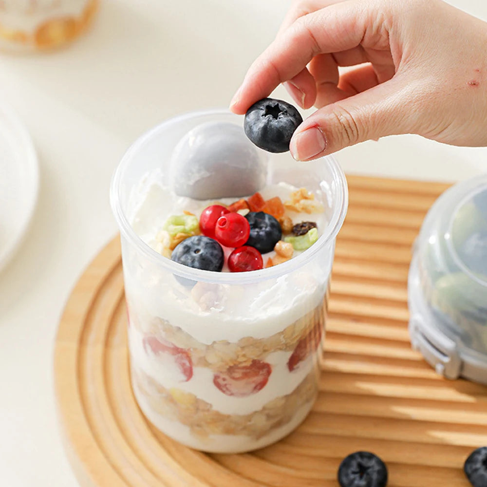 A hand is adding blueberries to a clear container of overnight oats on a wooden board.