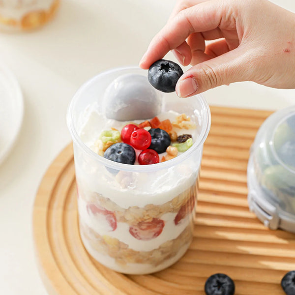 A hand is adding blueberries to a clear container of overnight oats on a wooden board.
