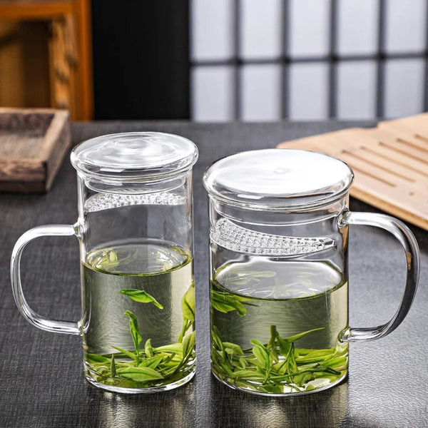 Klares Glas-Teebecher mit Henkel und Sichelfilter gefüllt mit grünen Blättern auf dunkler Oberfläche.