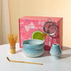 A pink box containing a matcha tea set with a ceramic bowl, whisk, and bamboo tools on a white surface.