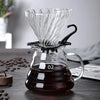 A clear glass manual pour over coffee dripper filled with dark brewed coffee sits on a dark wooden surface.