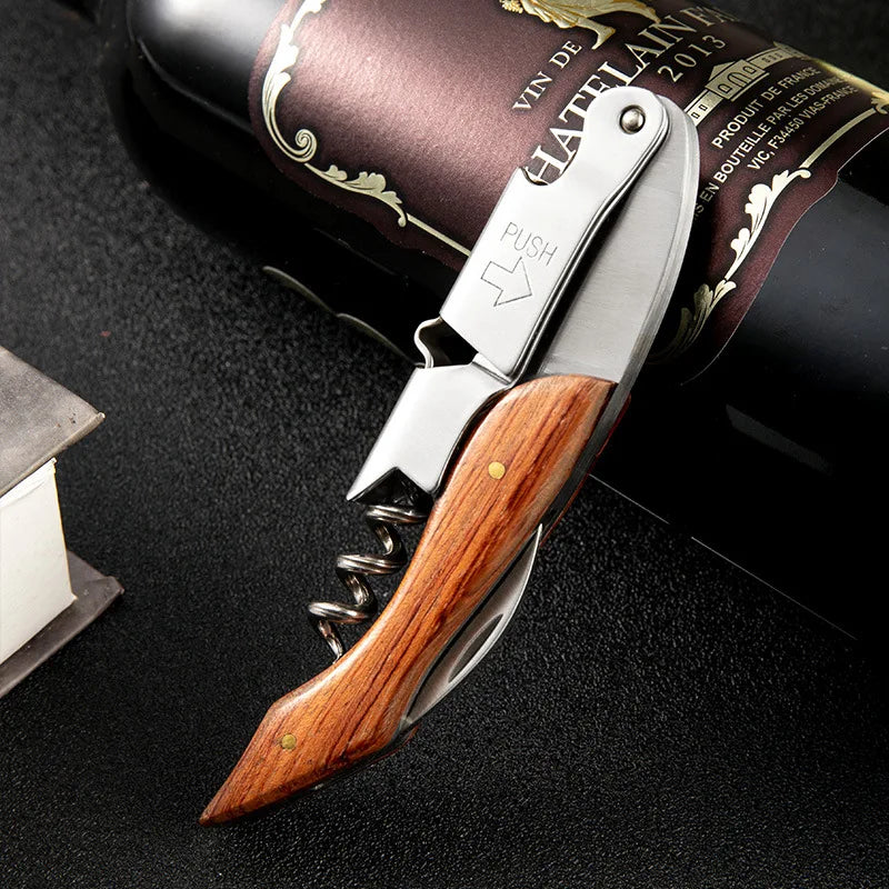 A silver double-hinged waiter's corkscrew with a wooden handle and foil cutter resting on a dark surface next to a wine bottl