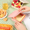 Hands are assembling a sandwich in a pink reusable microwave container on a green checkered mat.
