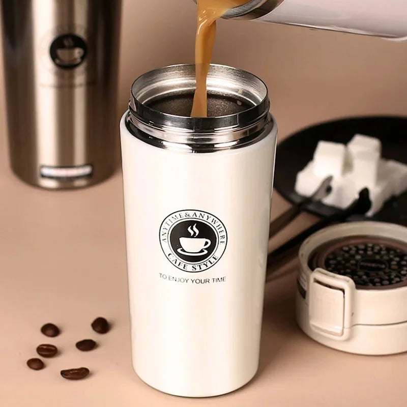 A white double-walled stainless steel thermal travel mug filled with light brown liquid, sitting next to coffee beans and a s
