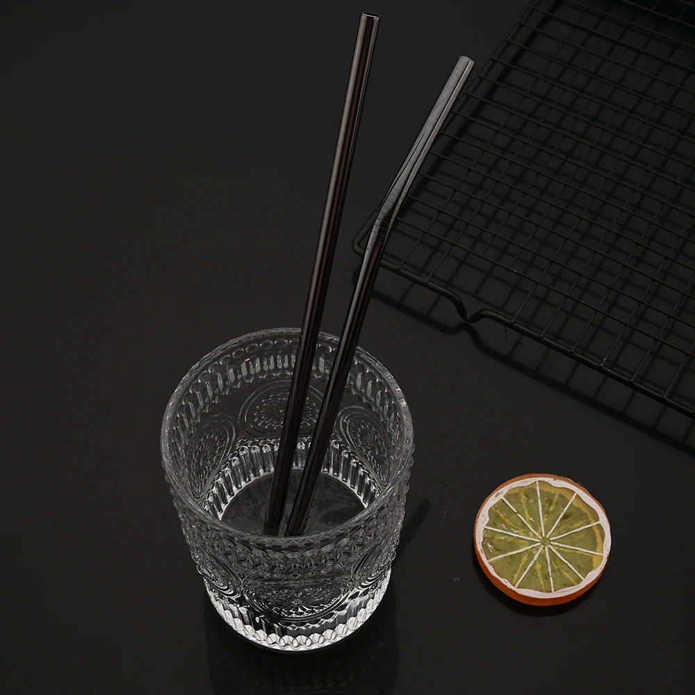 A glass filled with water and two black drinking straws rests on a black surface next to a bamboo mat and a slice of lime.