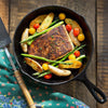 A cast iron frying pan filled with grilled fish, asparagus, and roasted potatoes on a wooden surface.