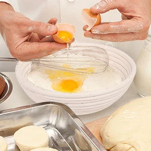 Hands are pouring egg yolk into a white bowl containing white powder, with a bread proofing basket and dough visible.