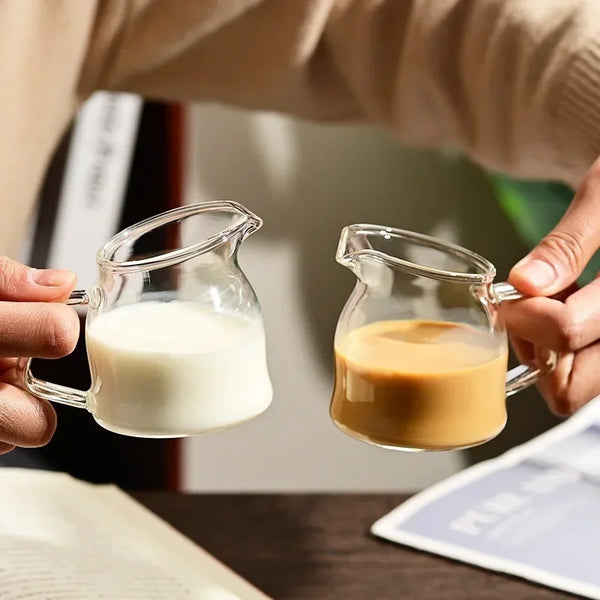A hand holds a clear glass serving cup filled with light-colored liquid, likely milk or coffee.