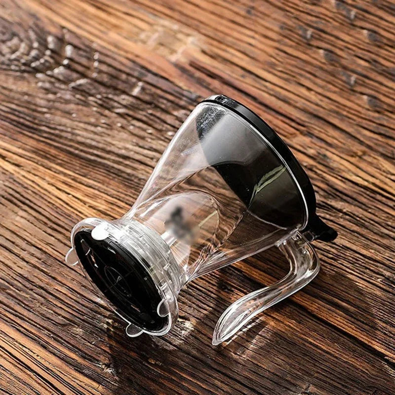 A clear glass pour over coffee dripper with a black handle resting on a wooden surface.