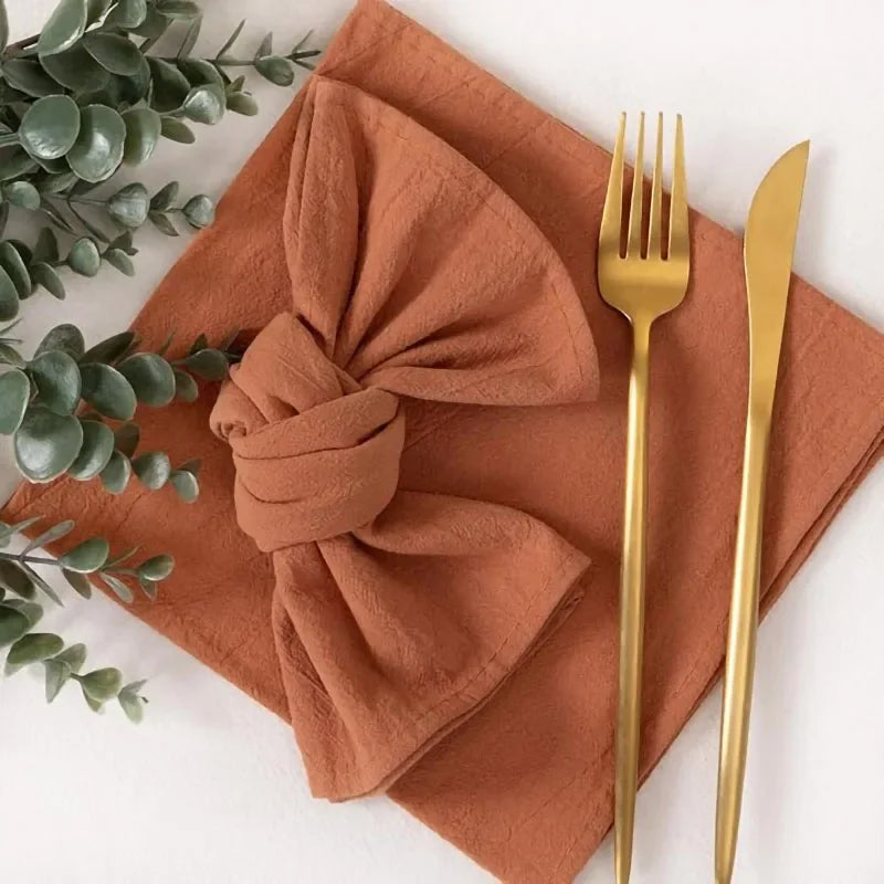 A set of reusable terracotta-colored cotton cloth napkins, a napkin bow, and gold cutlery on a white surface.