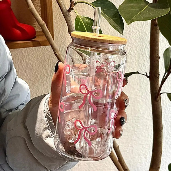 A hand holds a clear glass tumbler water bottle with pink ribbon designs against a background of green leaves and a tree bran