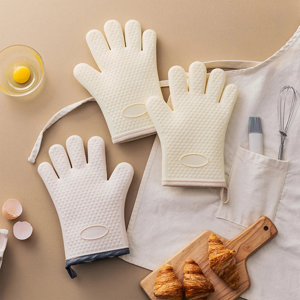 A set of white heat resistant silicone baking gloves resting on a light beige surface with a wooden cutting board and croissa