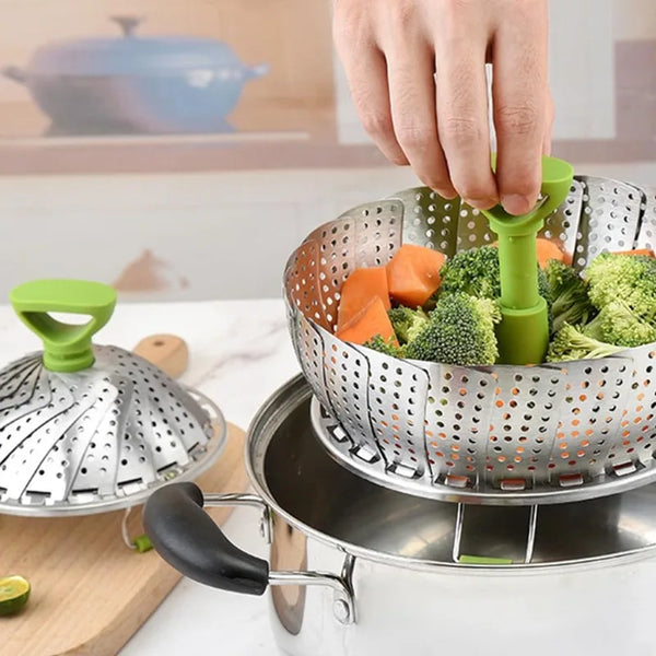 A hand is using a green handle to lift a stainless steel steamer basket filled with chopped vegetables.