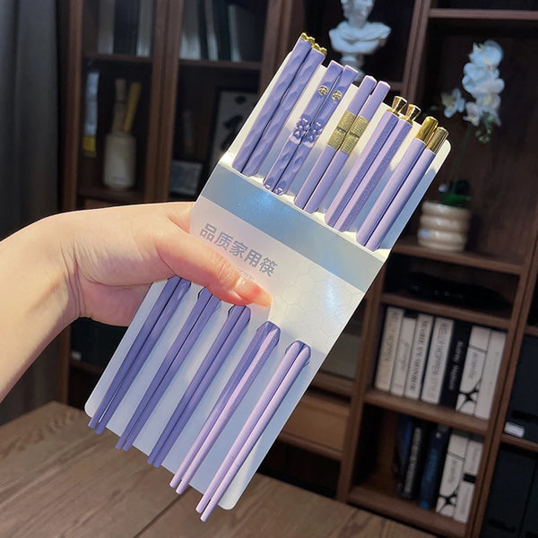 A hand holds a set of purple reusable anti-slip dining chopsticks against a wooden bookshelf background.