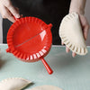 Hands are pressing dough into a red manual dumpling press, with another piece of dough nearby.