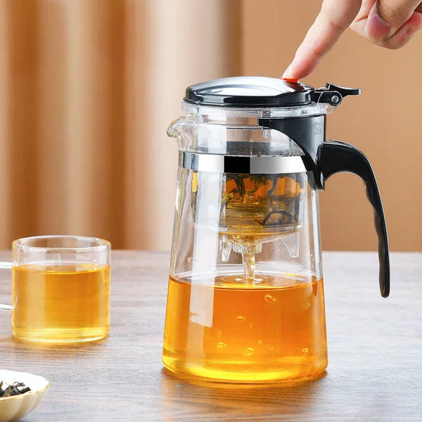 Eine Hand gießt goldene Flüssigkeit aus einer hitzebeständigen Glas-Tee-Kanne in eine Glas-Tasse.