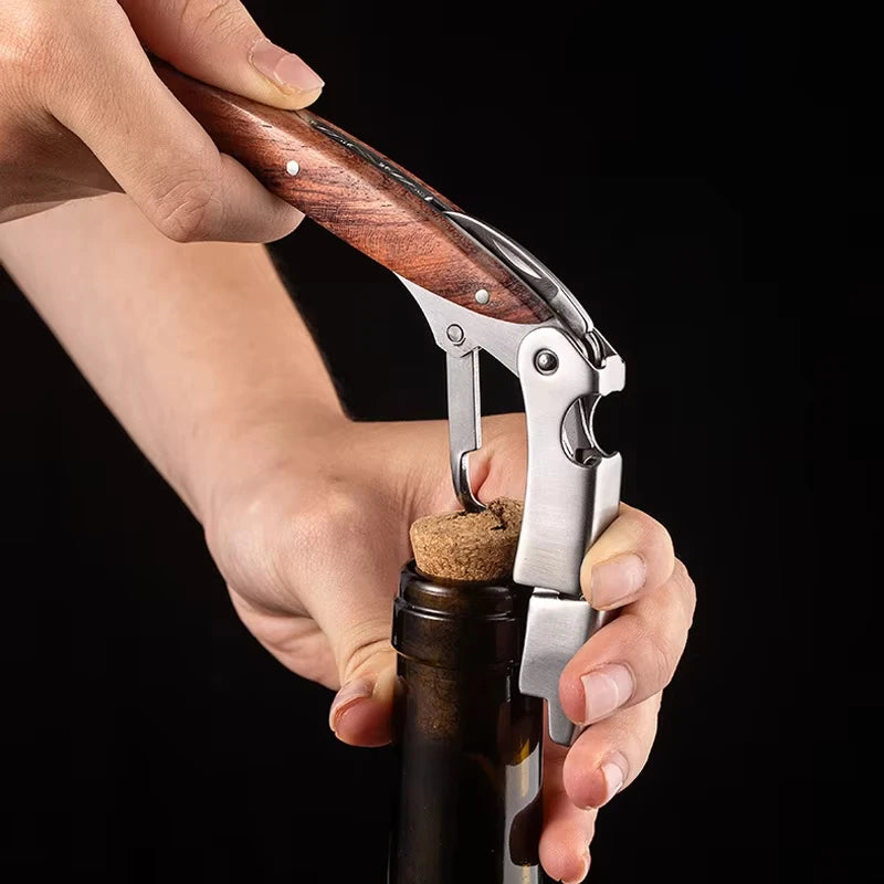 A hand holds a double-hinged waiter's corkscrew with a wooden handle and foil cutter over a bottle neck.