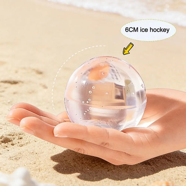 A hand holds a clear silicone ice hockey ball on a sandy beach.