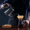 A person pours coffee into a glass using a metal pitcher and reusable cone coffee filters.