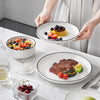 Hands are presenting a plate of grilled steak and vegetables on a white ceramic dinner plate.