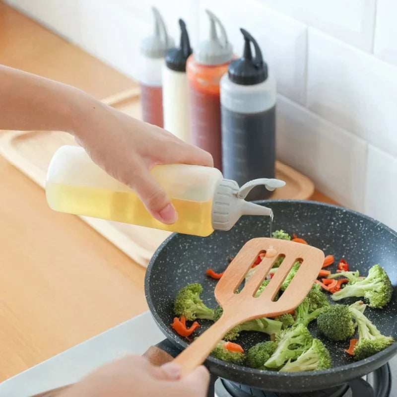 A hand pours yellow liquid seasoning from a squeeze dispenser bottle into a black frying pan filled with broccoli and carrots
