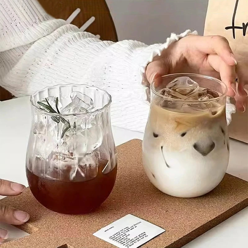 A glass of iced latte is placed on a wooden board next to a person's hand wearing a white sweater.