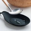 A black silicone spoon rest with a silver spoon and wooden handle, placed next to a woven basket.