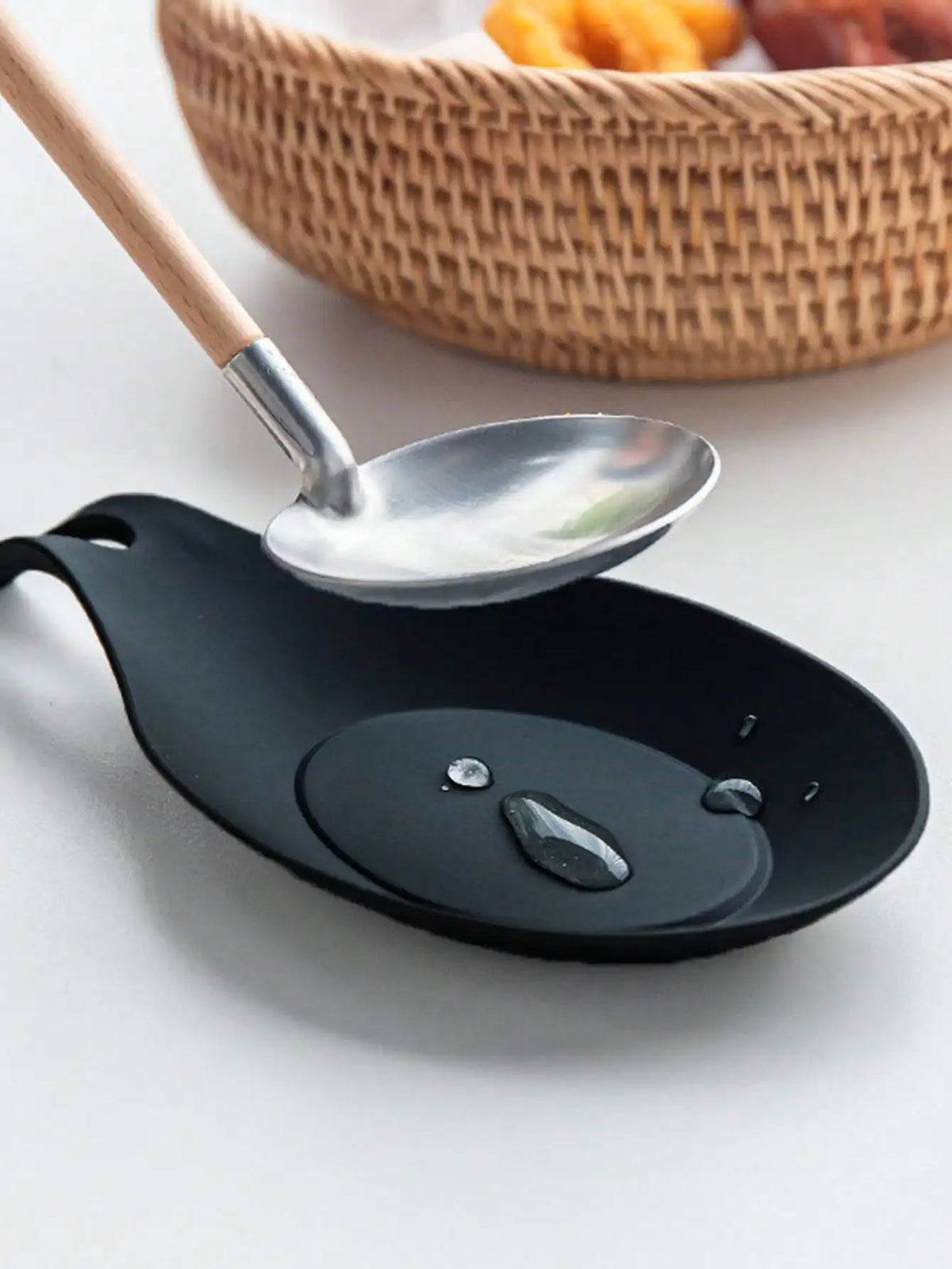 A black silicone spoon rest with a silver spoon and wooden handle, placed next to a woven basket.
