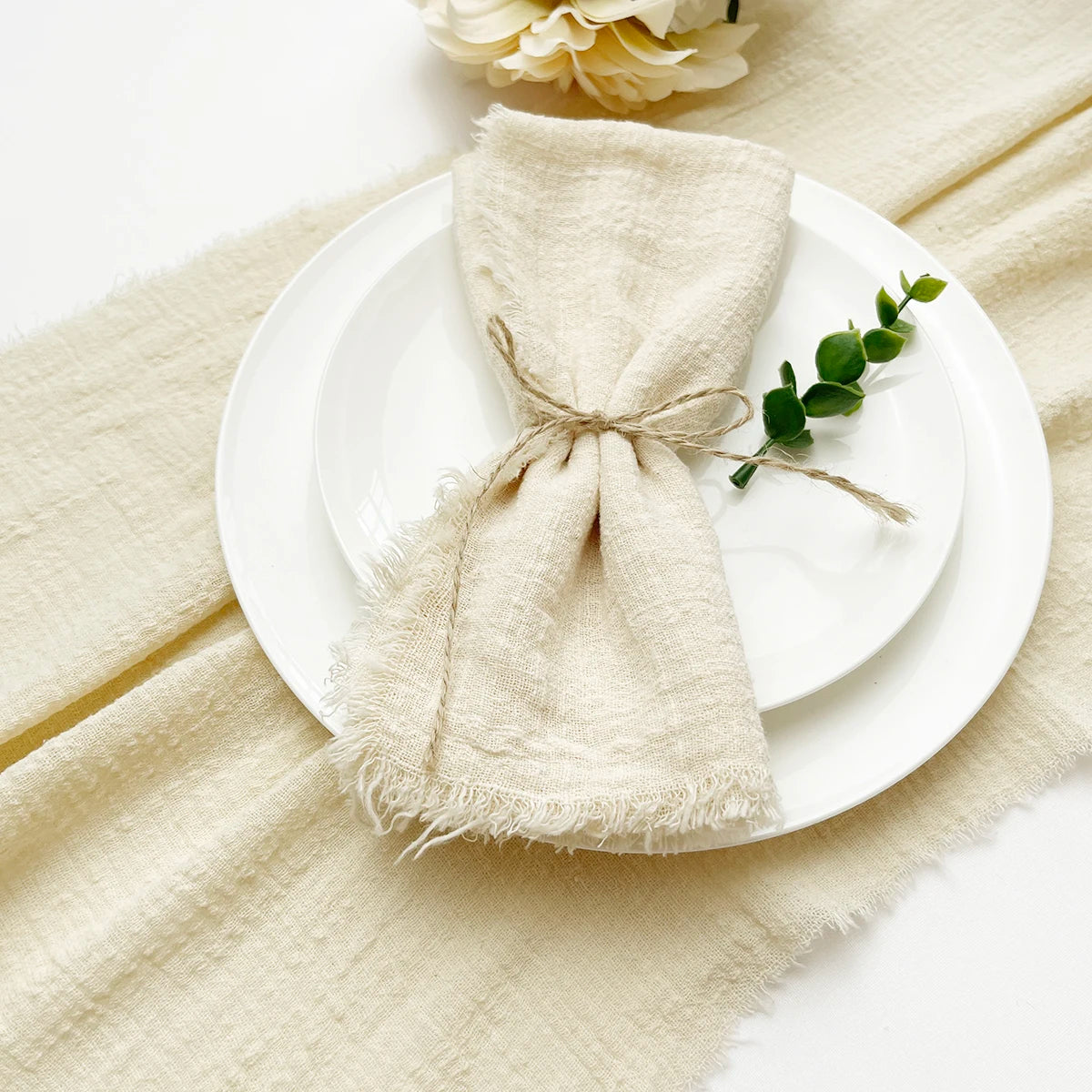Un paquete de servilletas de algodón reutilizables beige atadas con hilo, sobre platos blancos y un paño de lino color crema.