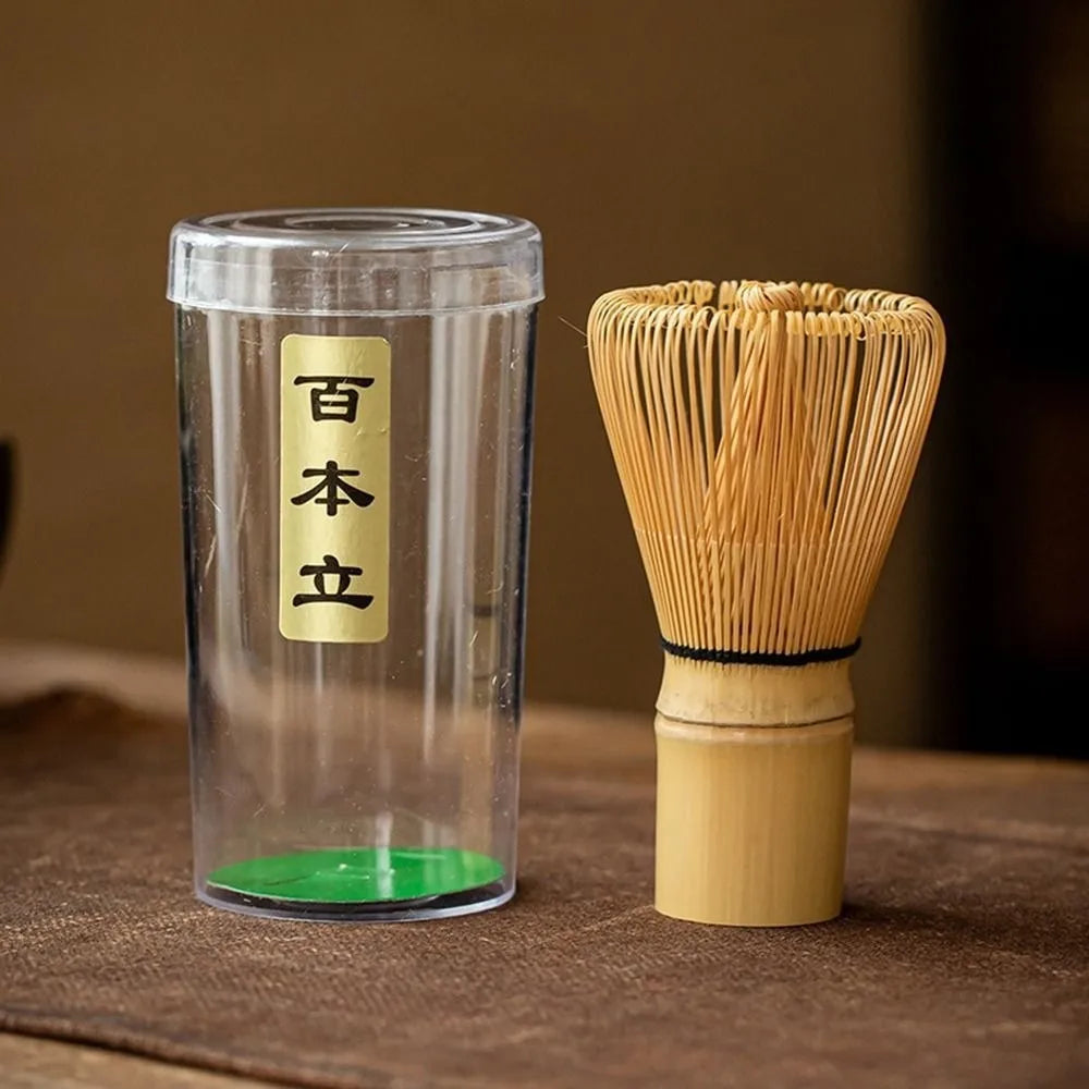 A glass container holds a bamboo matcha whisk and a green tea powder, representing a traditional Japanese matcha tea set.