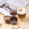 A glass of coffee with foam on a light brown background next to an open reusable coffee capsule container and a book.