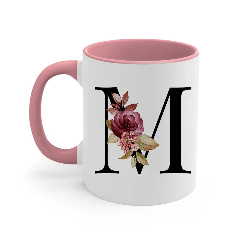 A pink ceramic coffee mug with a large black monogram featuring a rose and greenery design.