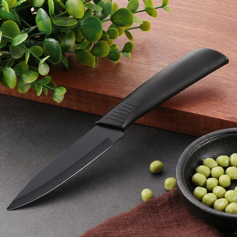 A black utility knife set with a dark handle rests on a dark surface next to green leaves and a bowl of peas.