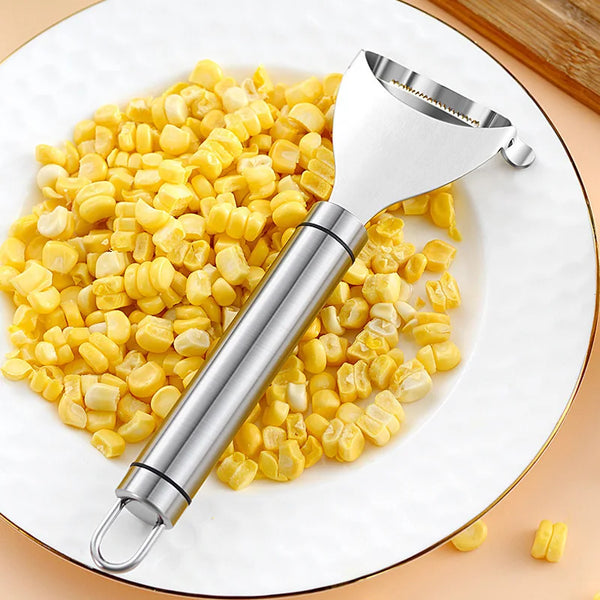 Silberner Edelstahl-Mais-Slicer und Schälerwerkzeug auf einer weißen Platte mit gelben Maiskörnern.