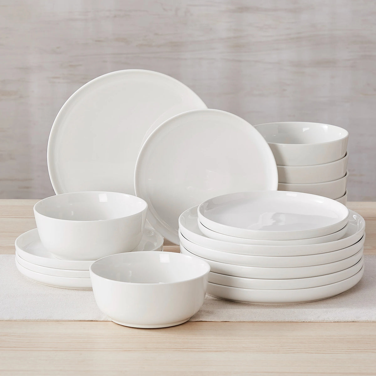 A white porcelain dinnerware set featuring plates and bowls stacked on a light wooden surface.