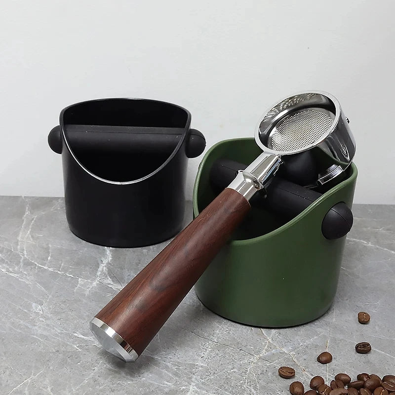 A black coffee knock box with a stainless steel filter and wooden handle sits next to coffee beans.