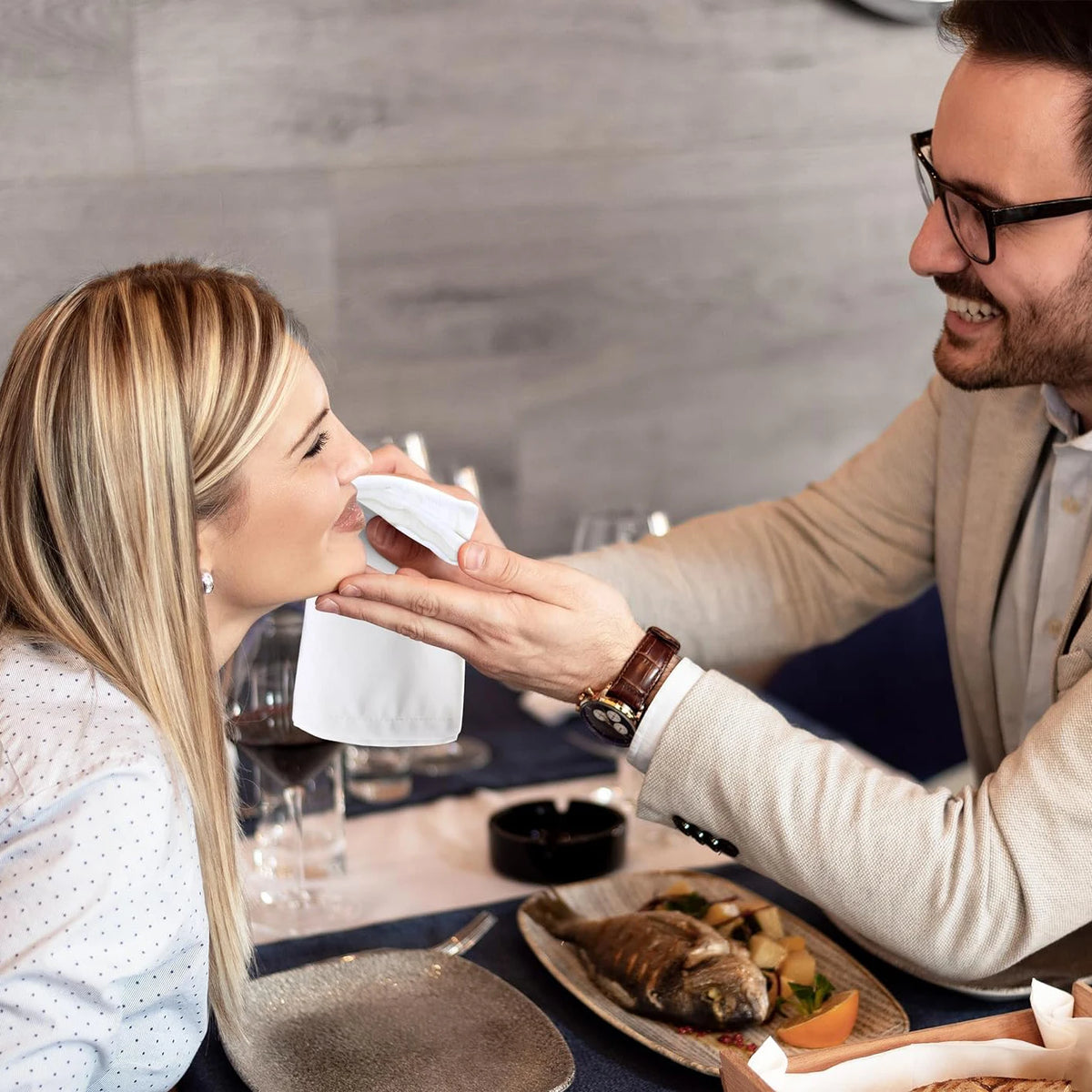 A woman is using a cloth napkin to wipe her mouth while dining, with food on a plate.