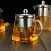 A clear glass teapot with a stainless steel infuser is filled with amber tea on a wooden surface.