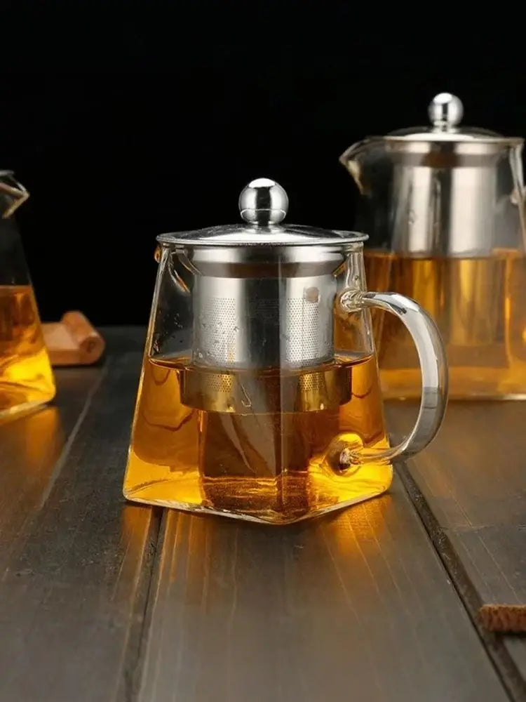A clear glass teapot with a stainless steel infuser is filled with amber tea on a wooden surface.