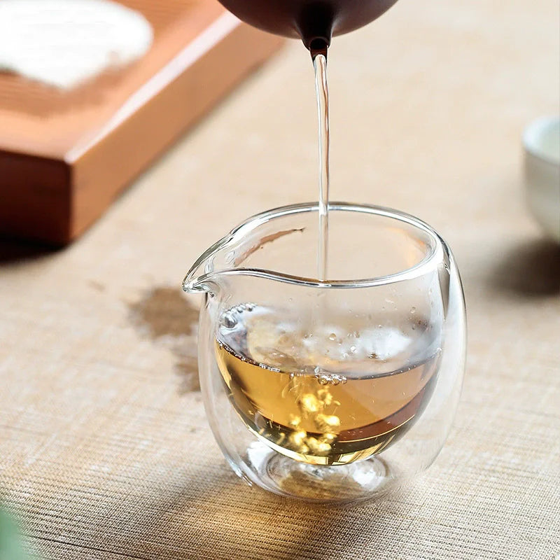 A glass tea pitcher is being filled with amber liquid from a dark spout.