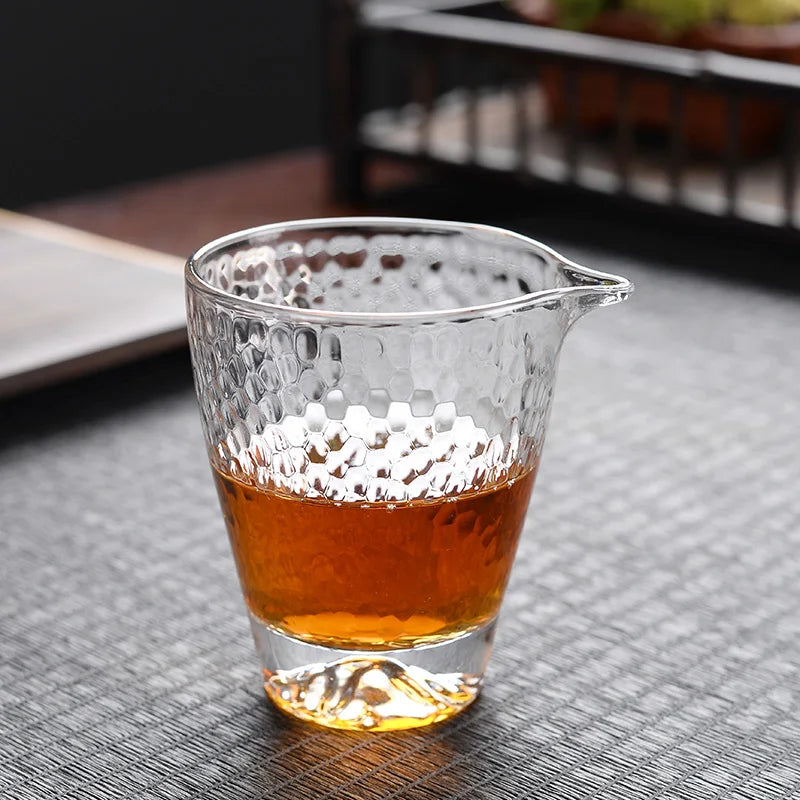 A clear, textured borosilicate glass tea cup is filled with amber-colored tea on a dark surface.