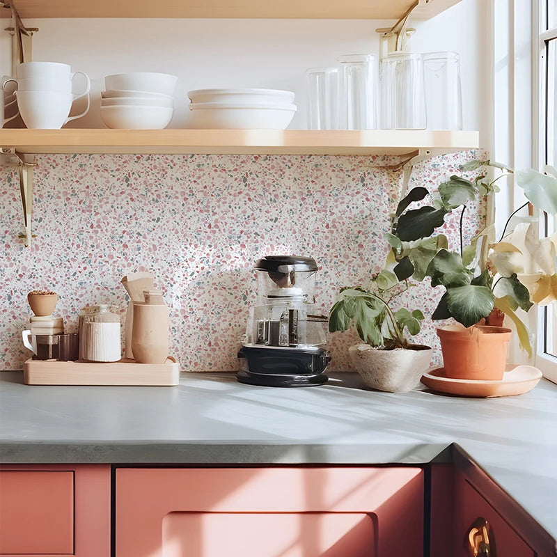 A kitchen shelf display featuring white ceramicware, glass jars, and potted greenery against a pink terrazzo patterned wall.