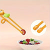 A pair of wooden kids training chopsticks with green grips is shown next to a bowl of fresh berries and fruit.